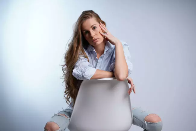 Hvit brunette dame med blå øyne sitter med bena spredt på en hvit stol i blå og hvit stripete skjorte og hullete jeans.